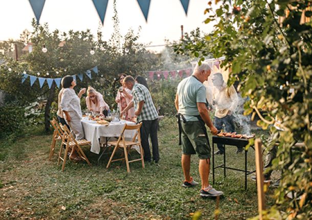 Mann grillt Fleisch im Garten, während die Familie den Tisch für das Mahl deckt.
