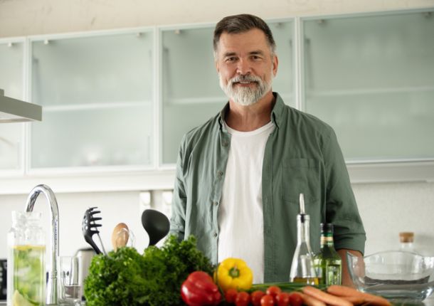 Lächelnder reifer Mann steht in der Küche, schaut in die Kamera und bereitet alleine zu Hause einen frischen Gemüsesalat zu.