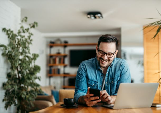 Porträt eines fröhlichen Mannes, der im Homeoffice ein Smartphone benutzt.