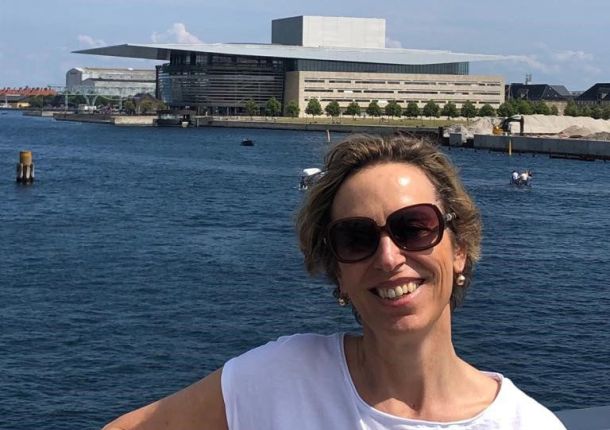 Frau lächelt am Wasser, mit einer Küste im Hintergrund.