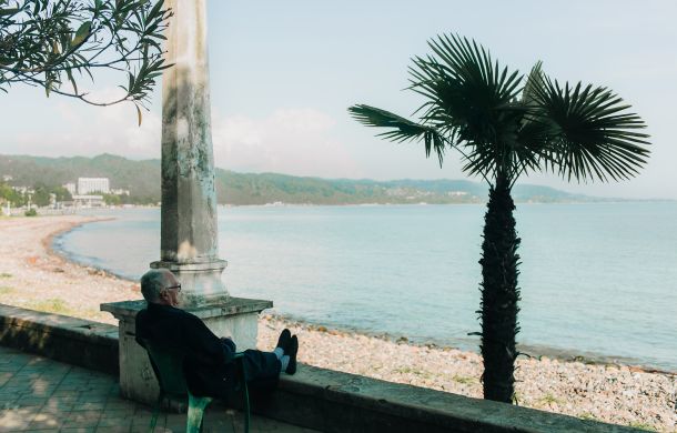 Älterer Mann sitzt entspannt auf einem Stuhl am Strand, genießt das Wetter und schaut aufs Meer während seines Urlaubs.